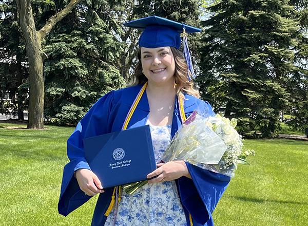 Cortney Rinehart in her graduation cap and gown, holding a bouquet of flowers and her diploma outside.