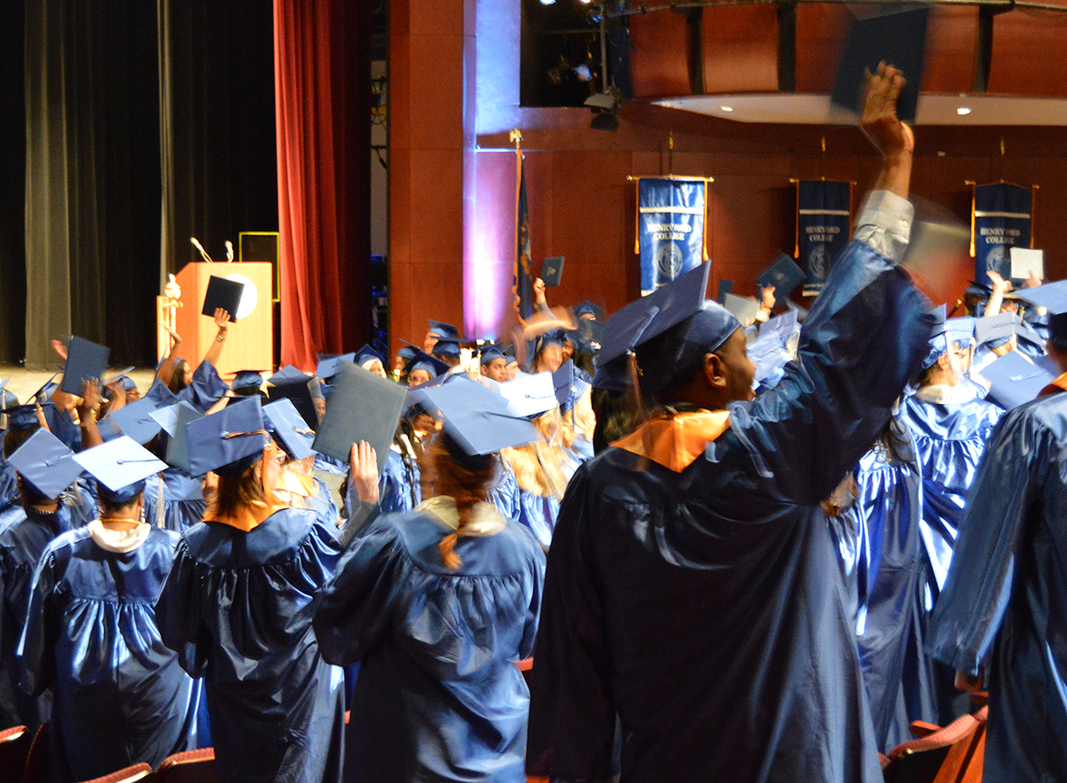 Numerous students wearing blue graduation gowns, some cheering and holding their caps up in the air