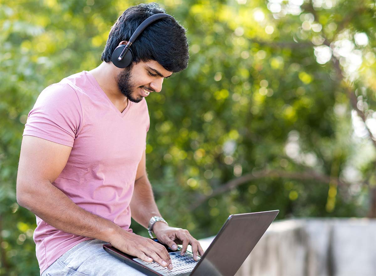 Student in pink shirt wearing headphones, smiling, while typing on laptop outside.