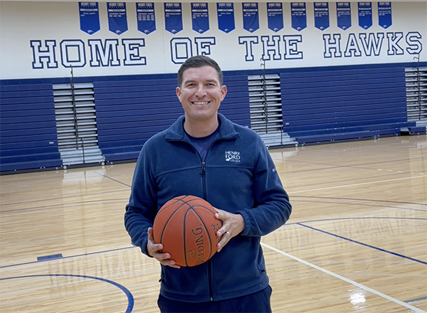 Russ Kavalhuna standing in a gym holding a basketball in front of "Home of the Hawks"