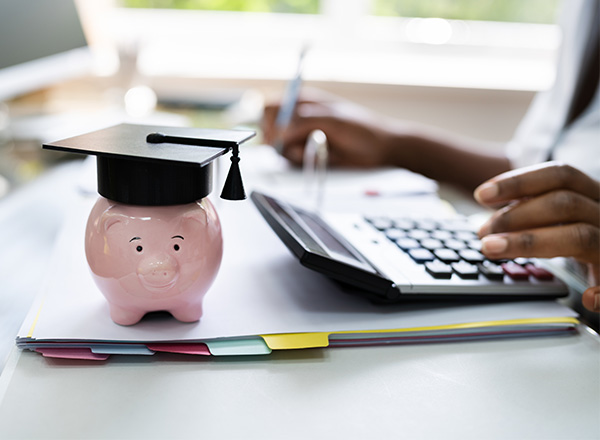 A person accounting next to a piggy bank wearing a graduation cap.