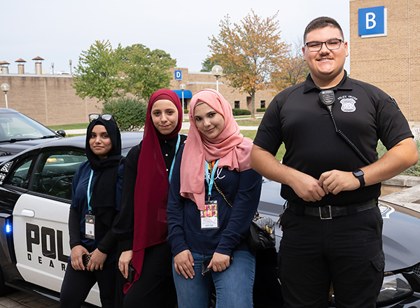 A police officer poses with three students in front of his cruiser. 