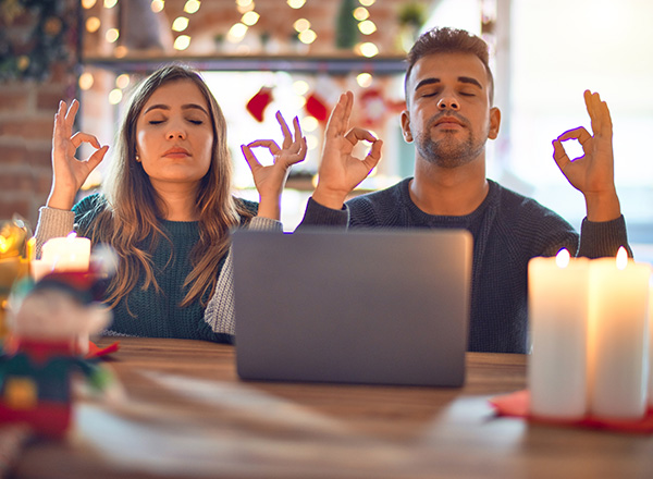 Young couple sitting using laptop around holiday decor at home with eyes closed doing meditation gesture with fingers. (Getty Images)