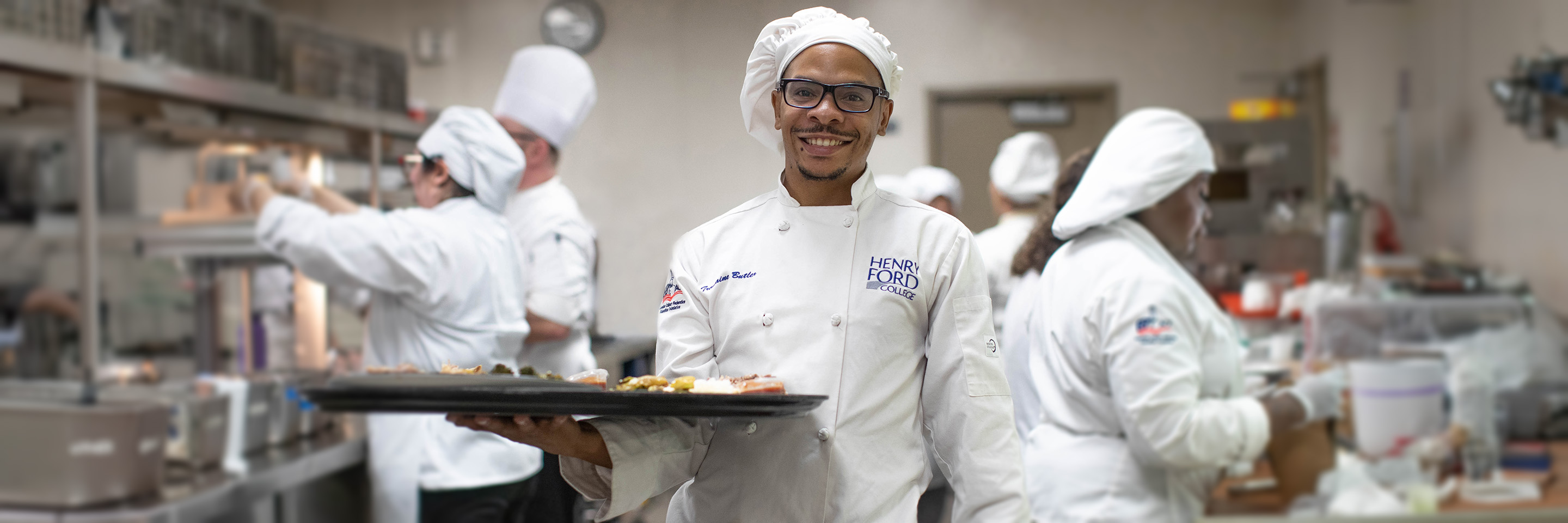 Culinary student proudly holding a tray of prepared food in one of HFC's kitchens, with other students cooking and prepping in the background