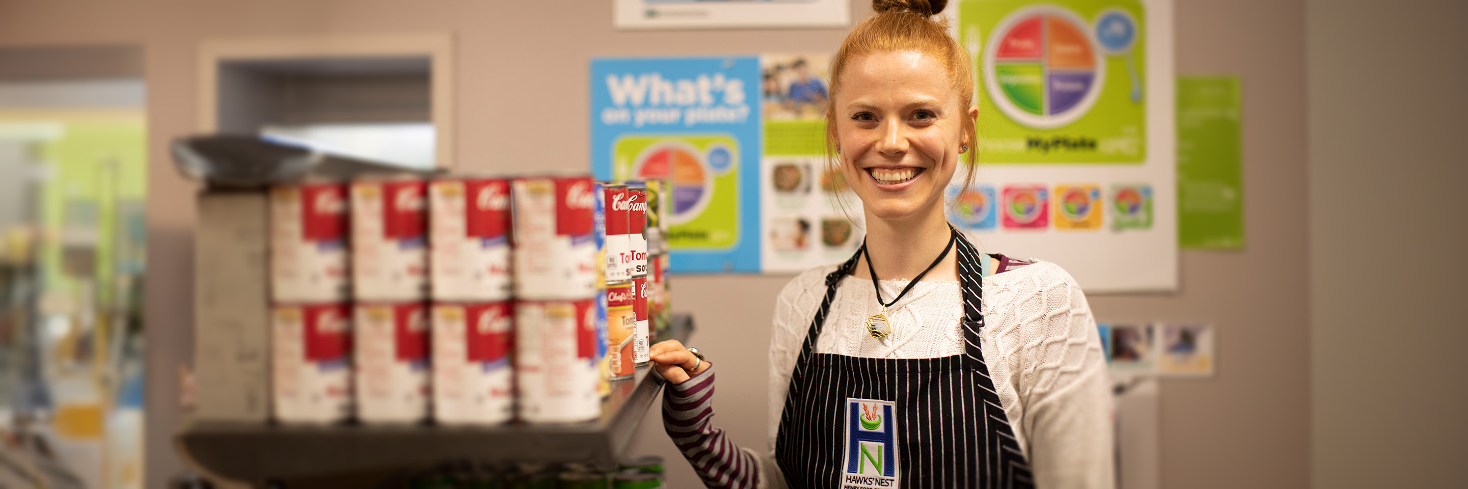 Student wearing an apron with the Hawks' Nest Food pantry logo, standing and smiling by a shelf of canned goods