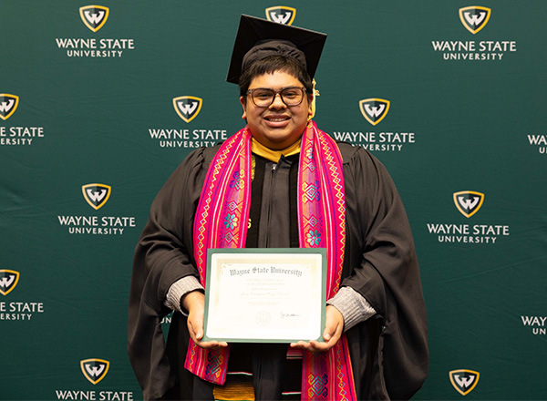 Jesus Cruz-Navarro is standing in front of a Wayne State University backdrop and proudly holds up his diploma from Wayne State University