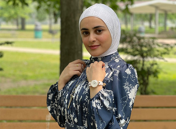 Zeinab Badreddine is wearing a white hijab and a navy and white floral dress, posing outdoors.