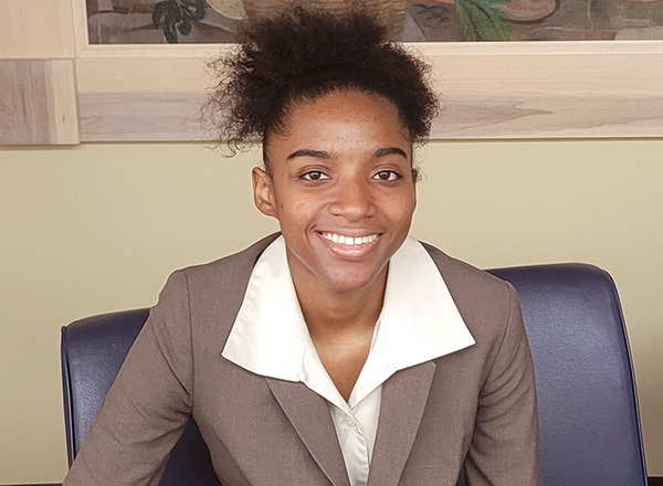 Lauryn Bolden, smiling and wearing a taupe business jacket with a white collared shirt, sitting on a blue chair.