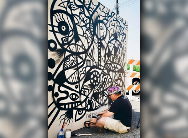 Daniel Cascardo is sitting on the ground and painting a wall outdoors.