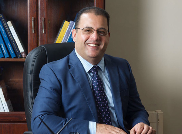 Dr. Naser Abumustafa is sitting at a desk with books in the background. He is wearing glasses and a navy suit and tie.