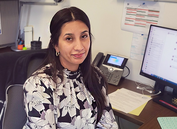 Amal Qureshi has long brown hair and dark eyes, wearing a black and white floral print blouse. She is sitting at her desk.