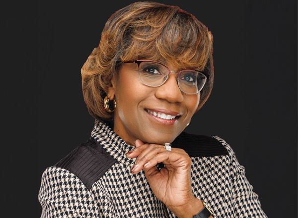 Linda Hannah has short caramel brown hair. She is wearing glasses and a houndstooth jacket in front of a dark background. 