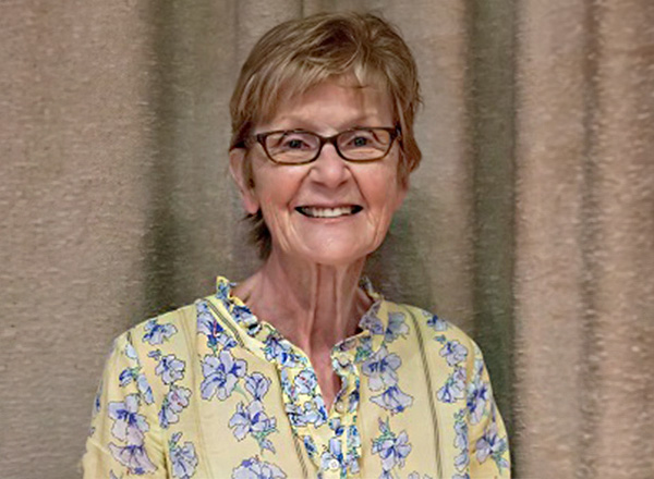 Mary Anne Hering has short light blonde hair, wearing glasses, and a yellow and blue floral blouse.