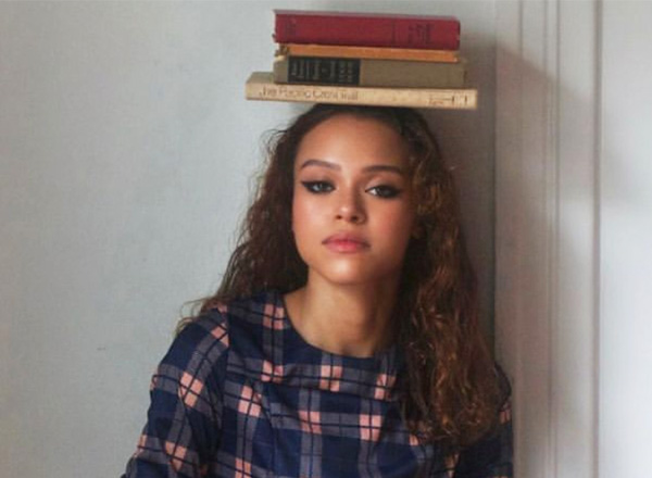 Diamond Arnold has long curly brown hair, wearing a plaid shirt, and balancing books on her head.