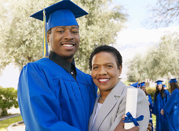 A graduate in blue cap and gown holding his diploma and hugging his mom.