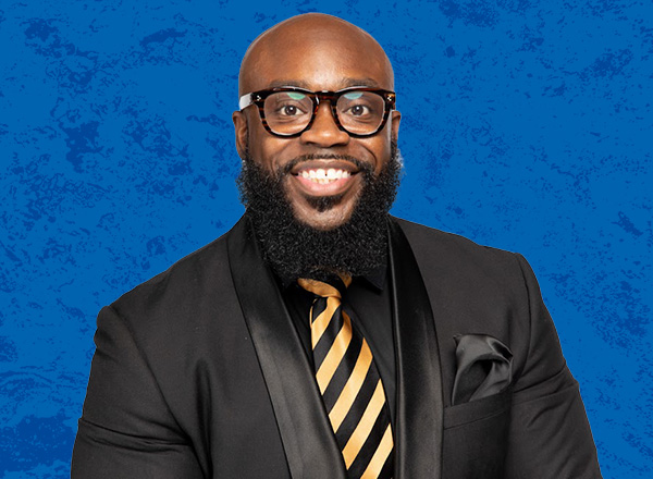 Keyon Clinton is wearing glasses, a dark suit with a striped tie, and has a beard. In front of a blue background.