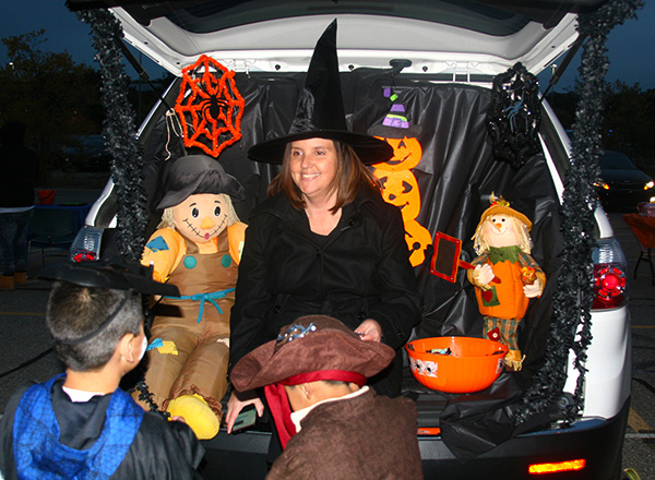 Woman dressed as a witch sitting in the trunk of her car handing out candy to trick or treaters.