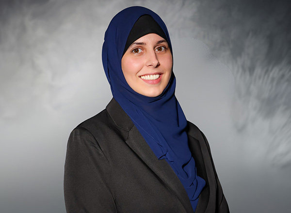 Shannon Wronski is wearing a blue hajib and black business jacket with a grey backdrop.