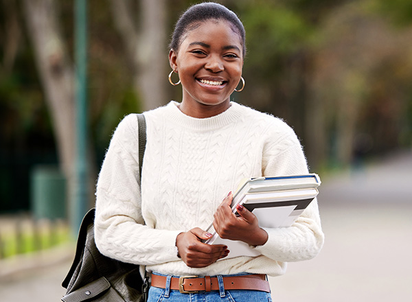 Smiling female student wearing a beige sweater, backpack, and holding books.