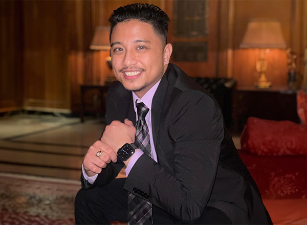 Marc Salamat sitting in a black suit with a lilac dress shirt and tie, smiling and adjusting watch. 