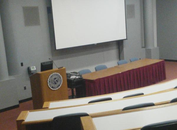 Photograph of the inside of Berry Auditorium, rows of auditorium seating and a large projector screen are visible, as well as a wooden podium next to a table with chairs in the front of the room in view of the "audience" 