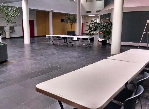 ASCC Atrium room L-111 pictured with tables and decorative plants; the room is large and has slate gray tiles, several white columns, and provides access to building exits and other rooms